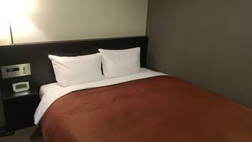 Una habitación de hotel con cama, almohadas blancas, colcha marrón, mesita de noche con reloj digital y una lámpara de pared.