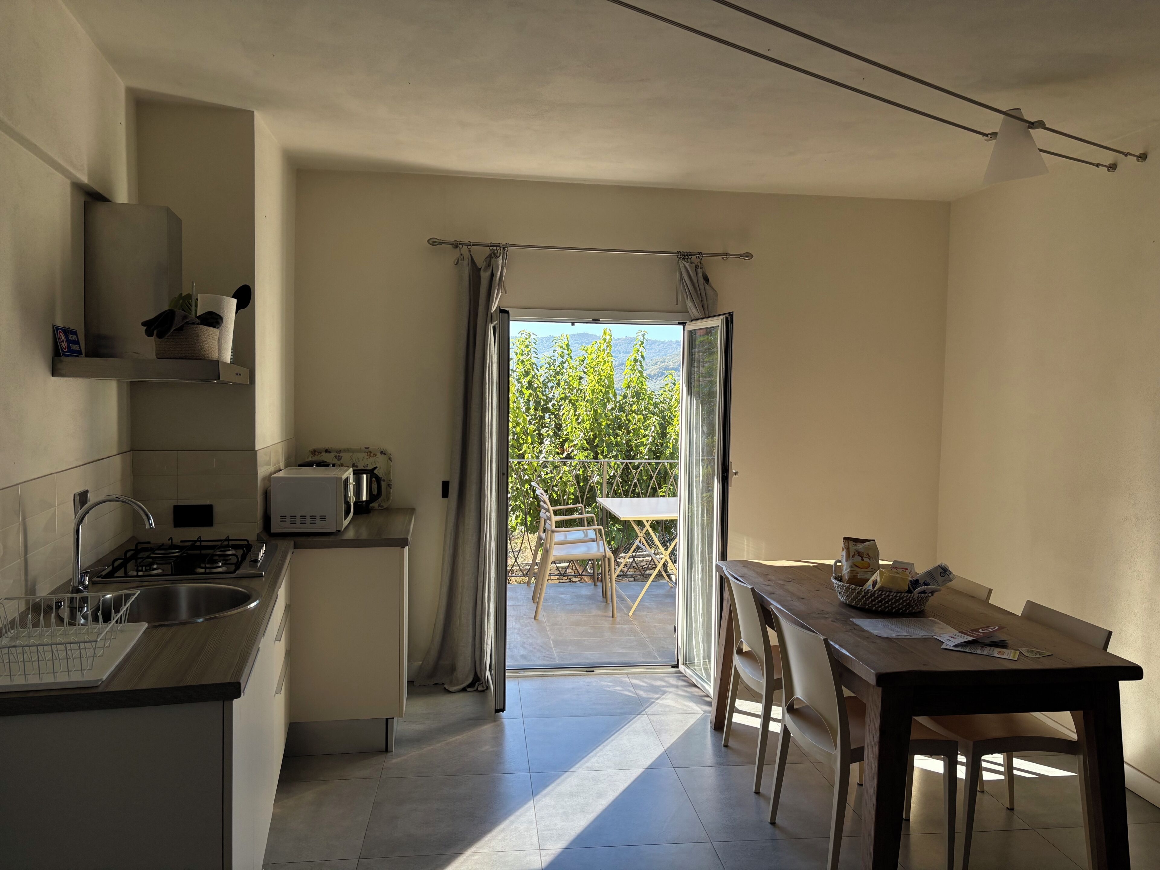 A compact kitchen with a gas stove, microwave, and sink. A dining area with a table and chairs is visible through an open door leading to a balcony with a view of trees.