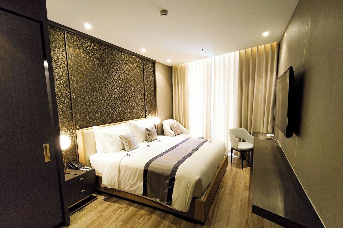 A modern hotel room with a large bed, a desk with a lamp, a chair, a flat-screen TV mounted on the wall, and a decorative wall panel.
