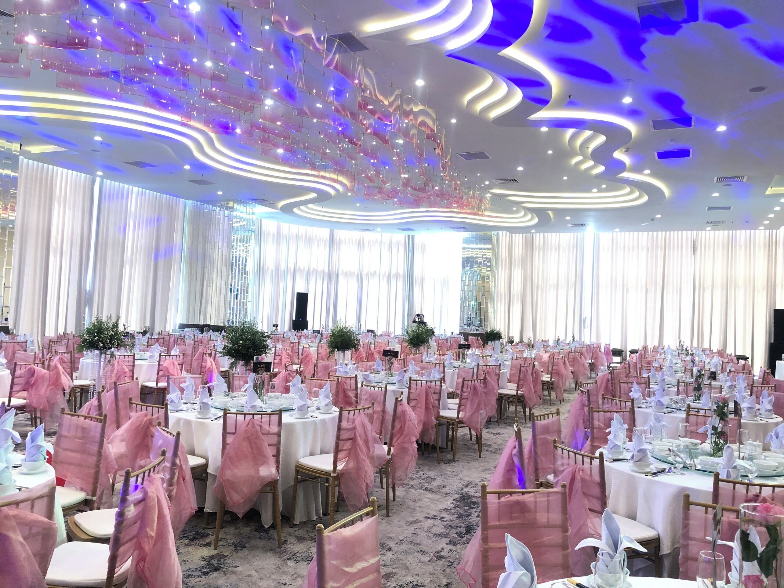 A banquet hall with round tables, pink chair covers, and a modern ceiling design.
