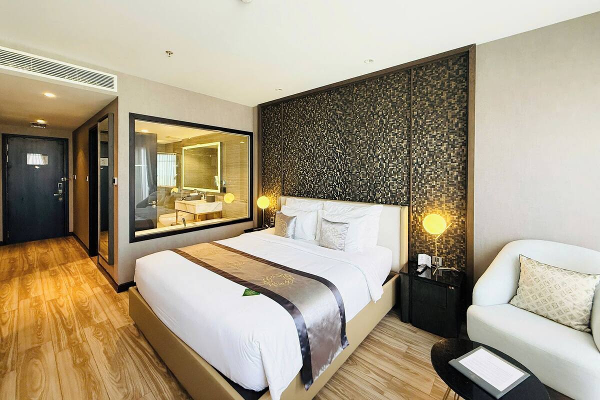 A hotel room with a large bed, a sitting area with a chair, a large mirror, a wall-mounted light, and a small table.