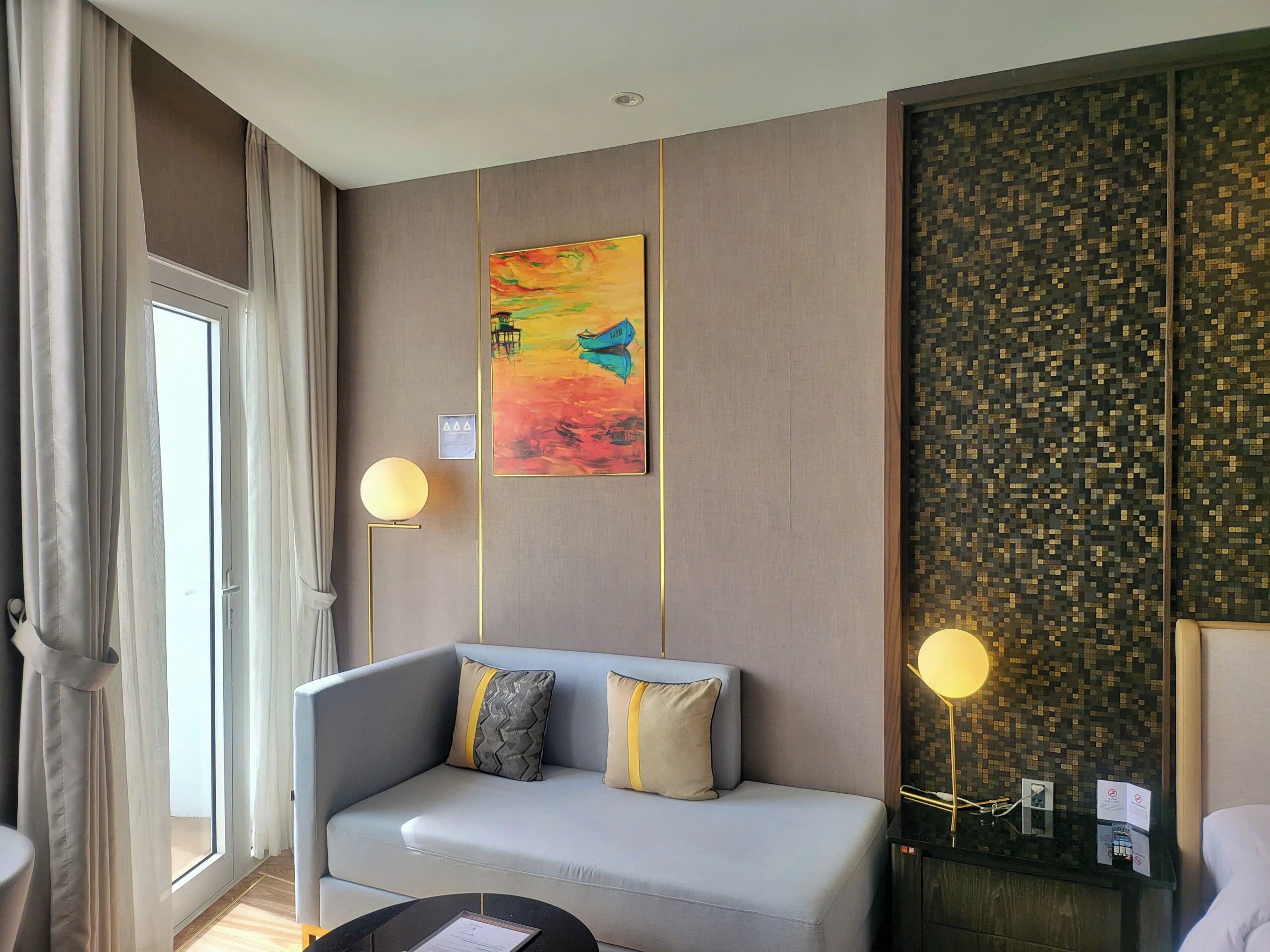 A modern hotel room with a grey sofa, two pillows, a painting, a wall with a pixelated pattern, and two wall-mounted lamps.