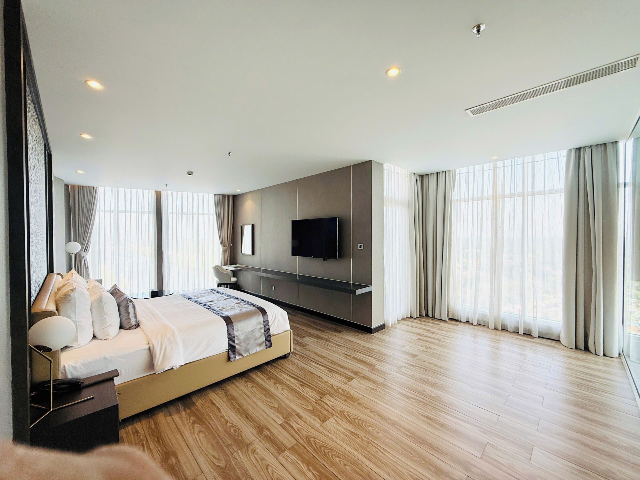 Quarto de hotel moderno com uma cama grande, uma escrivaninha, televisão de tela plana e janelas amplas com cortinas.