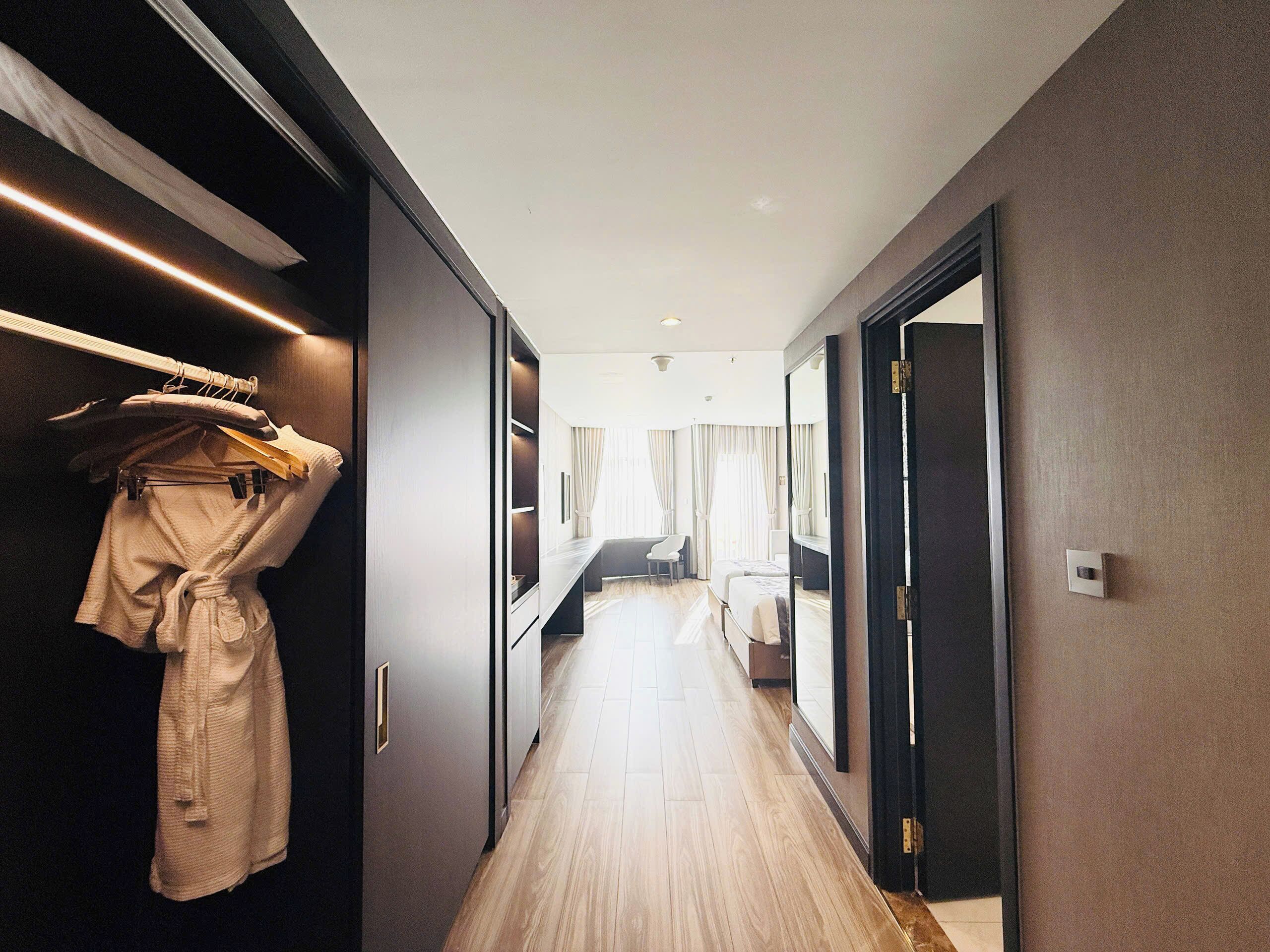 A hotel hallway with a wooden floor, a bed, a desk, and a wardrobe with clothes hanging.
