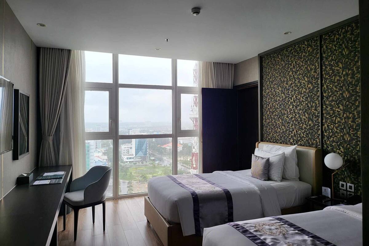 A hotel room with a large window, a desk, two beds, and a view of the cityscape.