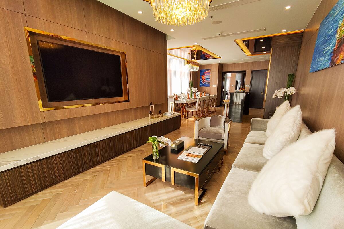 A modern living room with a large flat-screen TV, a sofa set, a coffee table, and a dining area in the background.