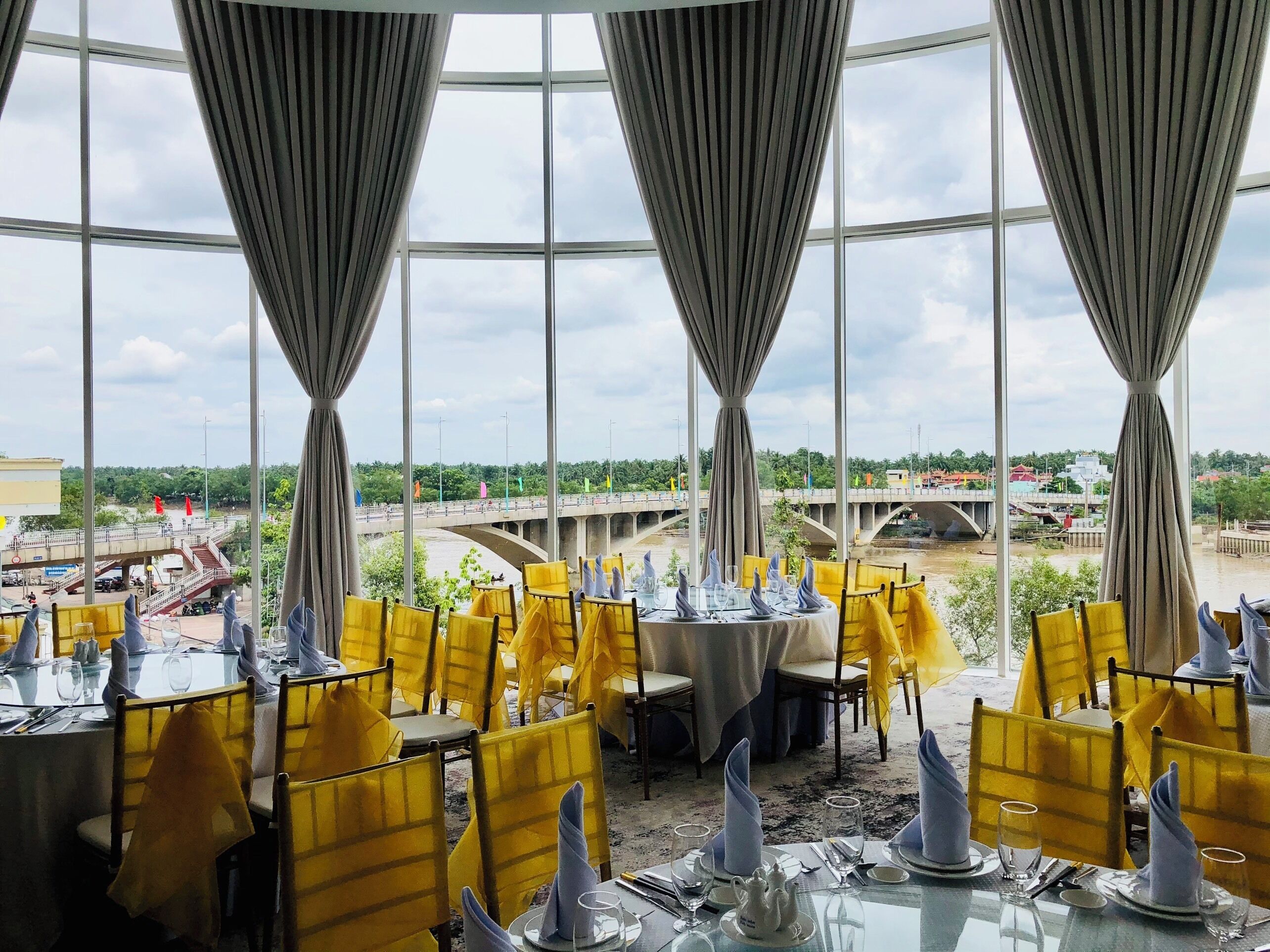 A restaurant with tables set for dining, large windows, and a view of a bridge and greenery.