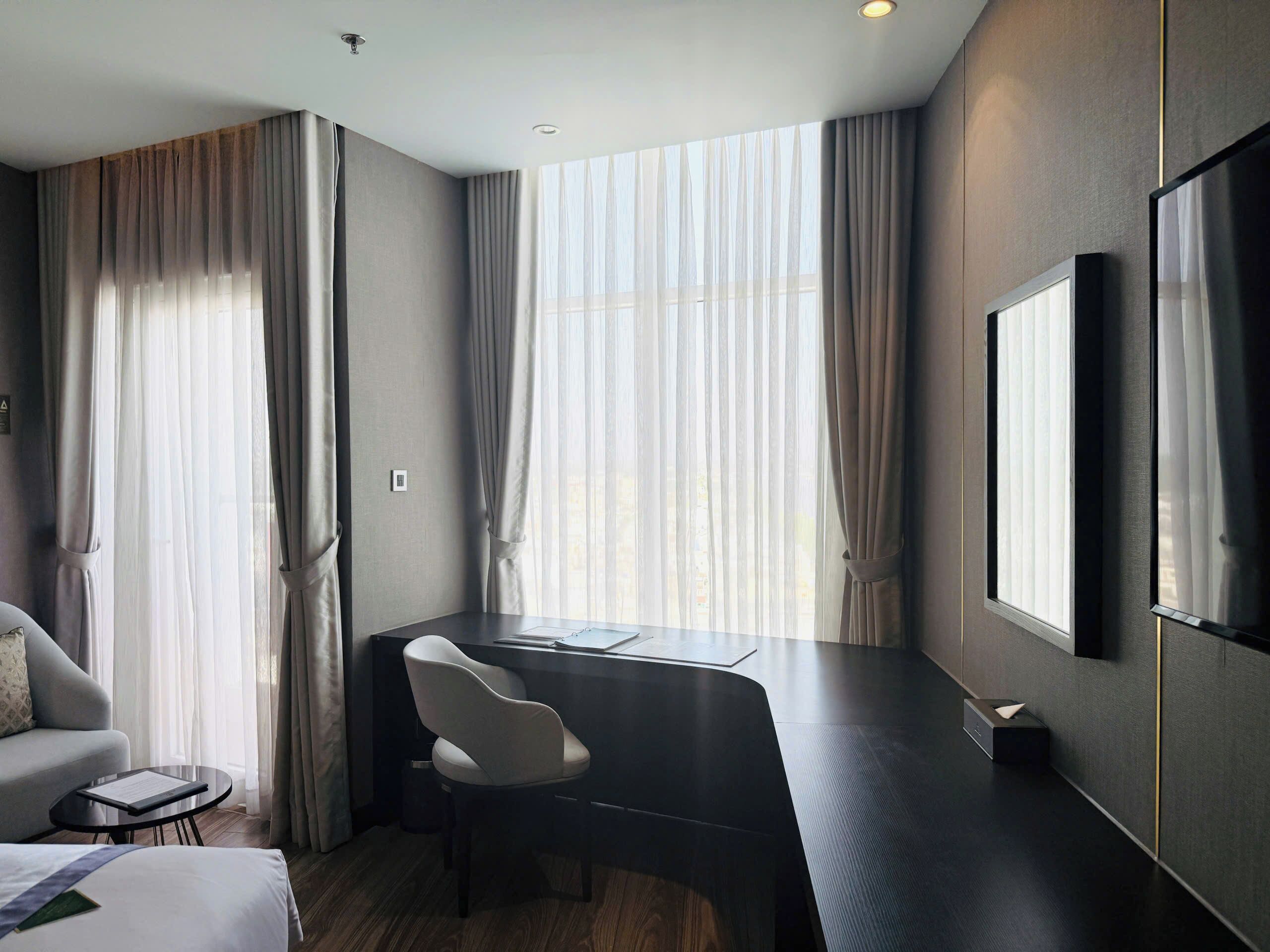 Ein Hotelzimmer mit Schreibtisch, Stuhl, Bett und großen Fenstern mit Vorhängen.