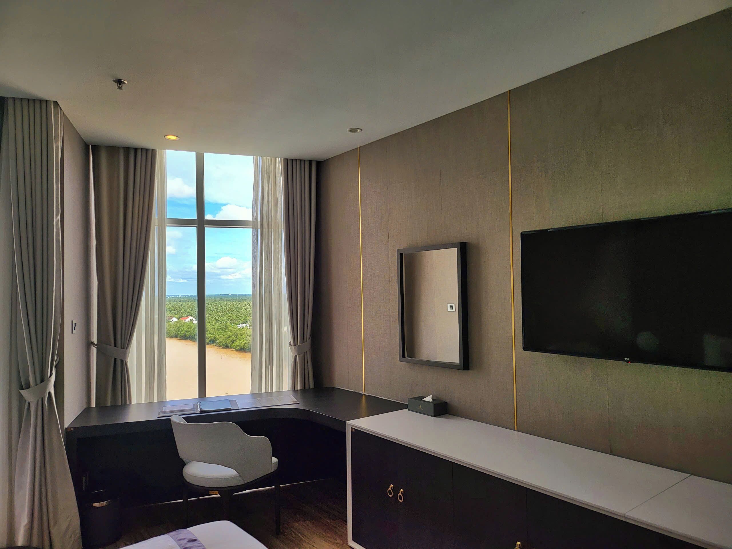 Habitación de hotel moderna con un ventanal grande, un escritorio, una silla, un televisor y un armario.