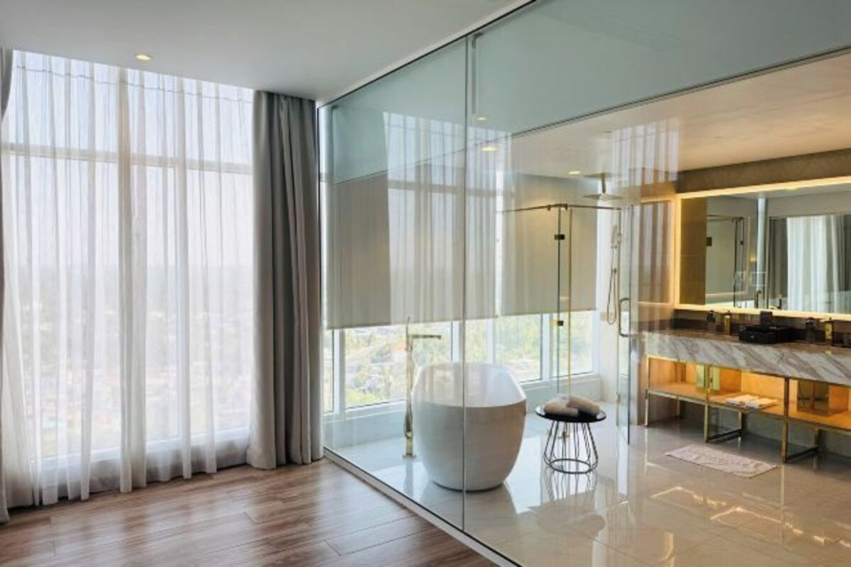 Quarto de hotel moderno com uma janela ampla, divisória de vidro, banheiro em mármore e uma cadeira branca arredondada.
