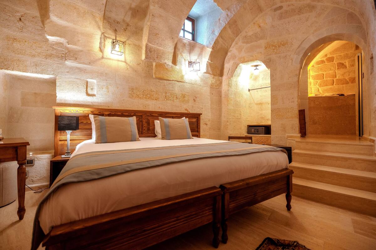 A bedroom with a large bed, wooden headboard, and stone walls. There are two bedside tables with lamps and a window above the bed.