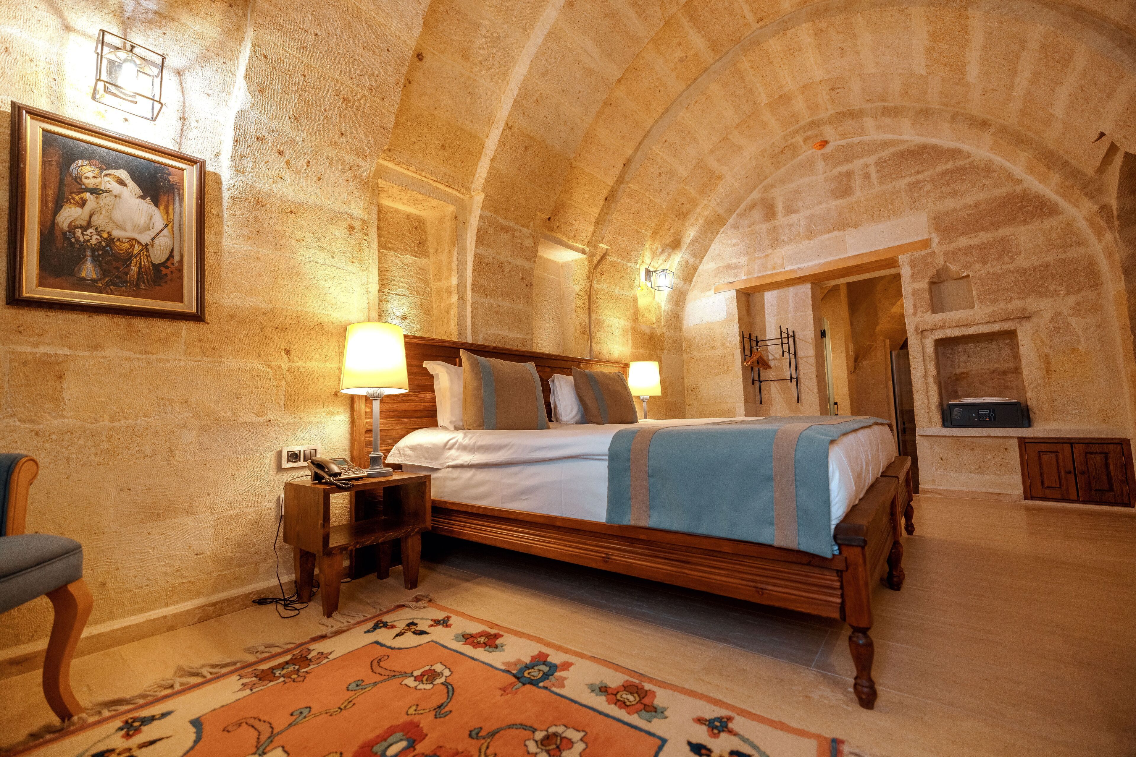 A bedroom with a wooden bed, two bedside lamps, a nightstand, a chair, a rug, and a stone wall.