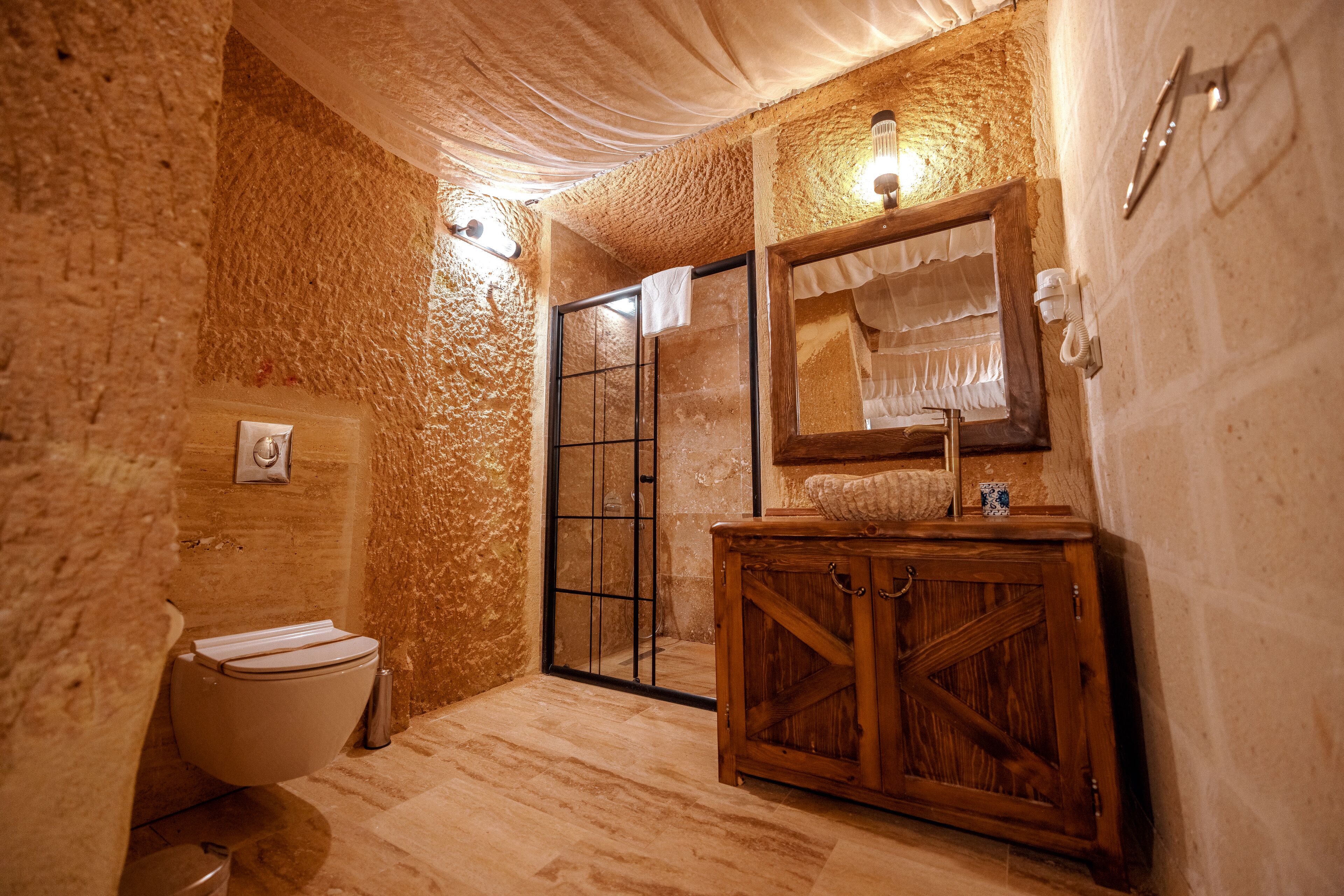 Une salle de bain avec un mur en pierre rustique, un meuble en bois, une cabine de douche en verre et des toilettes blanches.