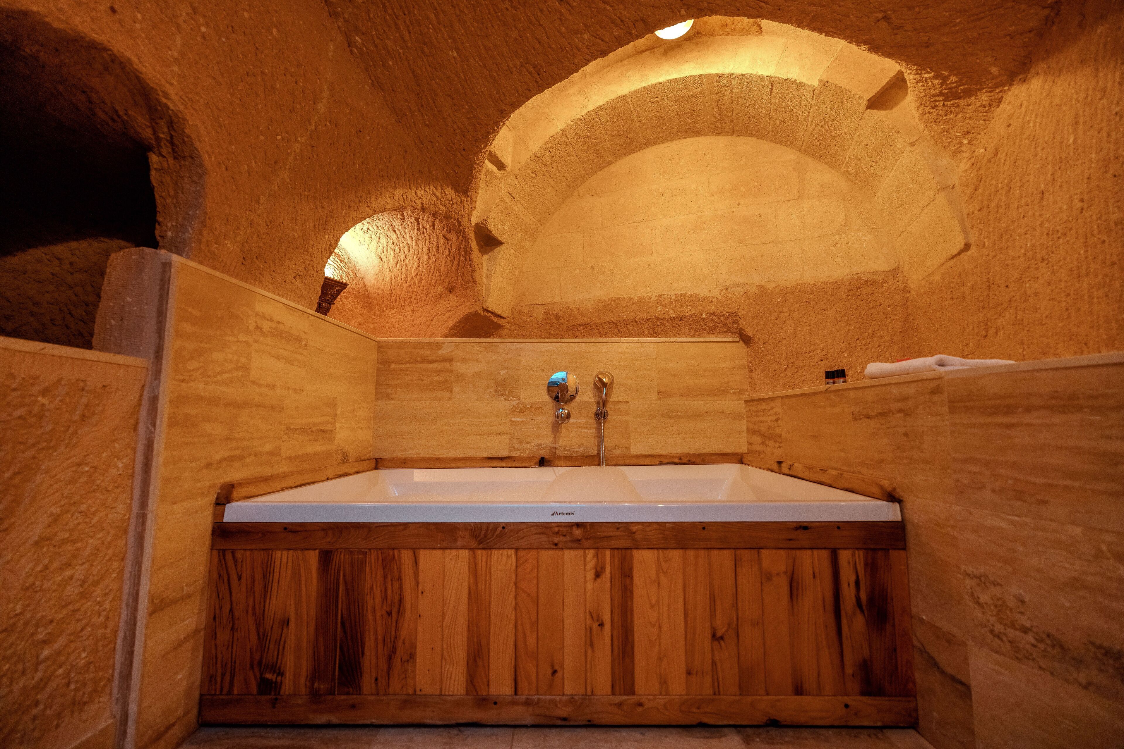 A wooden bathtub in a stone-walled room with a wooden door and a towel on the wall.