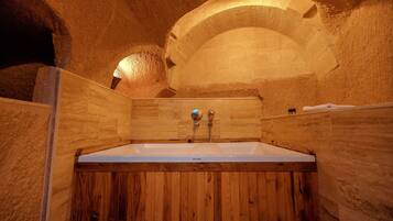 A wooden bathtub in a stone-walled room with a wooden door and a towel on the wall.