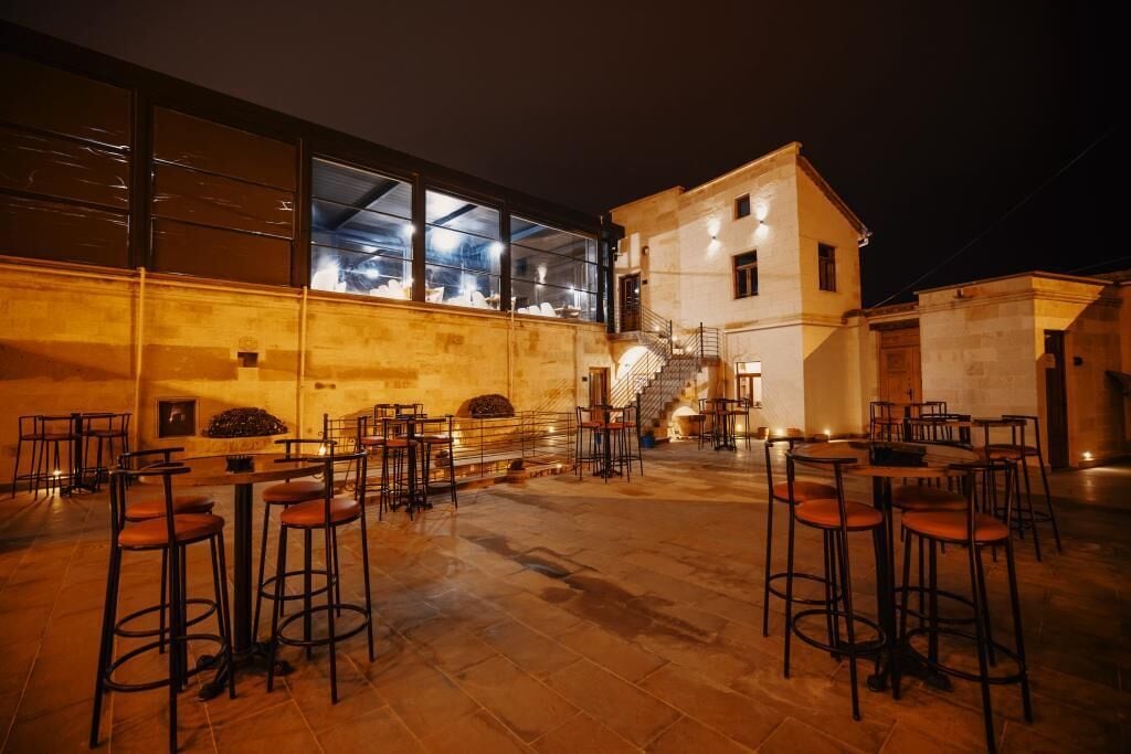 Uma área de bar ao ar livre com várias mesas e bancos, uma escadaria que leva a um nível superior e um edifício com grandes janelas ao fundo.
