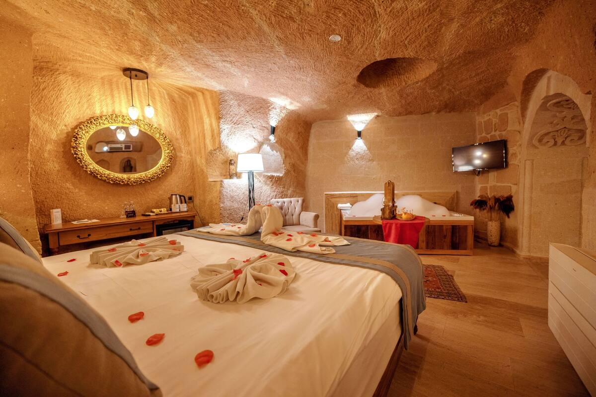 A hotel room with a wooden ceiling, a round mirror, a bed with rose petals, a bedside table, a TV, and a red tablecloth.
