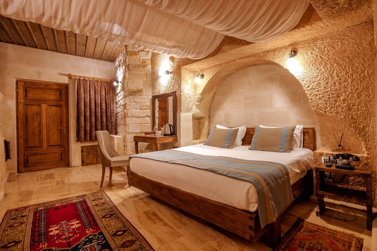 A bedroom with a large bed, wooden headboard, stone walls, a wooden desk, and a chair.