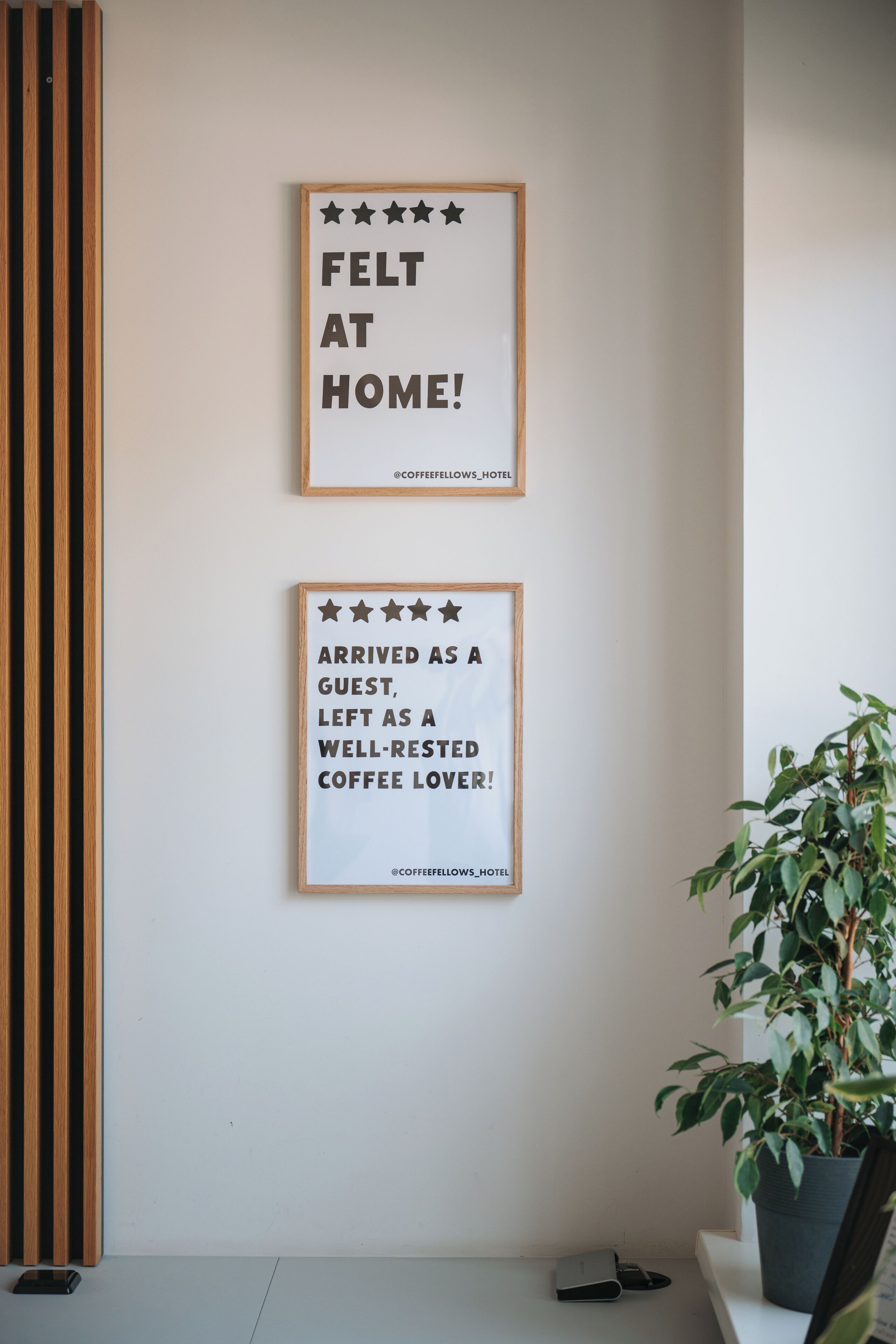 Two framed signs on a wall, one reads 'FELT AT HOME!' and the other 'ARRIVED AS A GUEST, LEFT AS A WELL-RESTED COFFEE LOVER!' with a social media handle '@COFFEEFELLOWS_HOTEL' at the bottom.