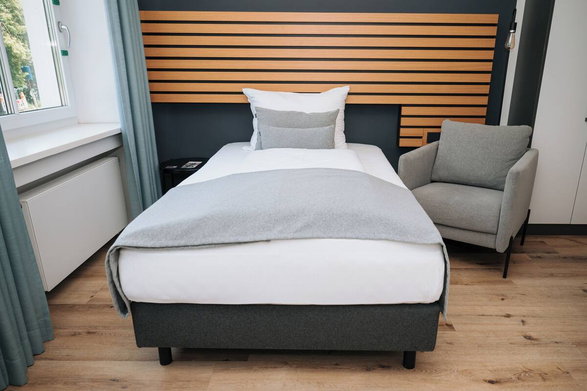 Un dormitorio moderno con cama, ropa de cama gris y blanca, un sillón gris y cabecero de madera.
