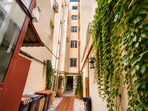 Un callejón estrecho con un edificio a un lado y una ventana al otro. Se ven botes de basura y una planta en maceta.