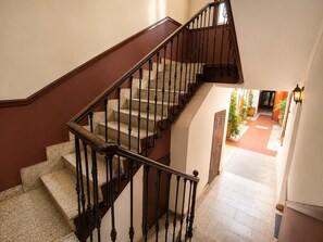 Una escalera con barandilla de madera y balaustradas negras, que lleva a un rellano con una puerta y vistas a un patio con plantas en macetas.