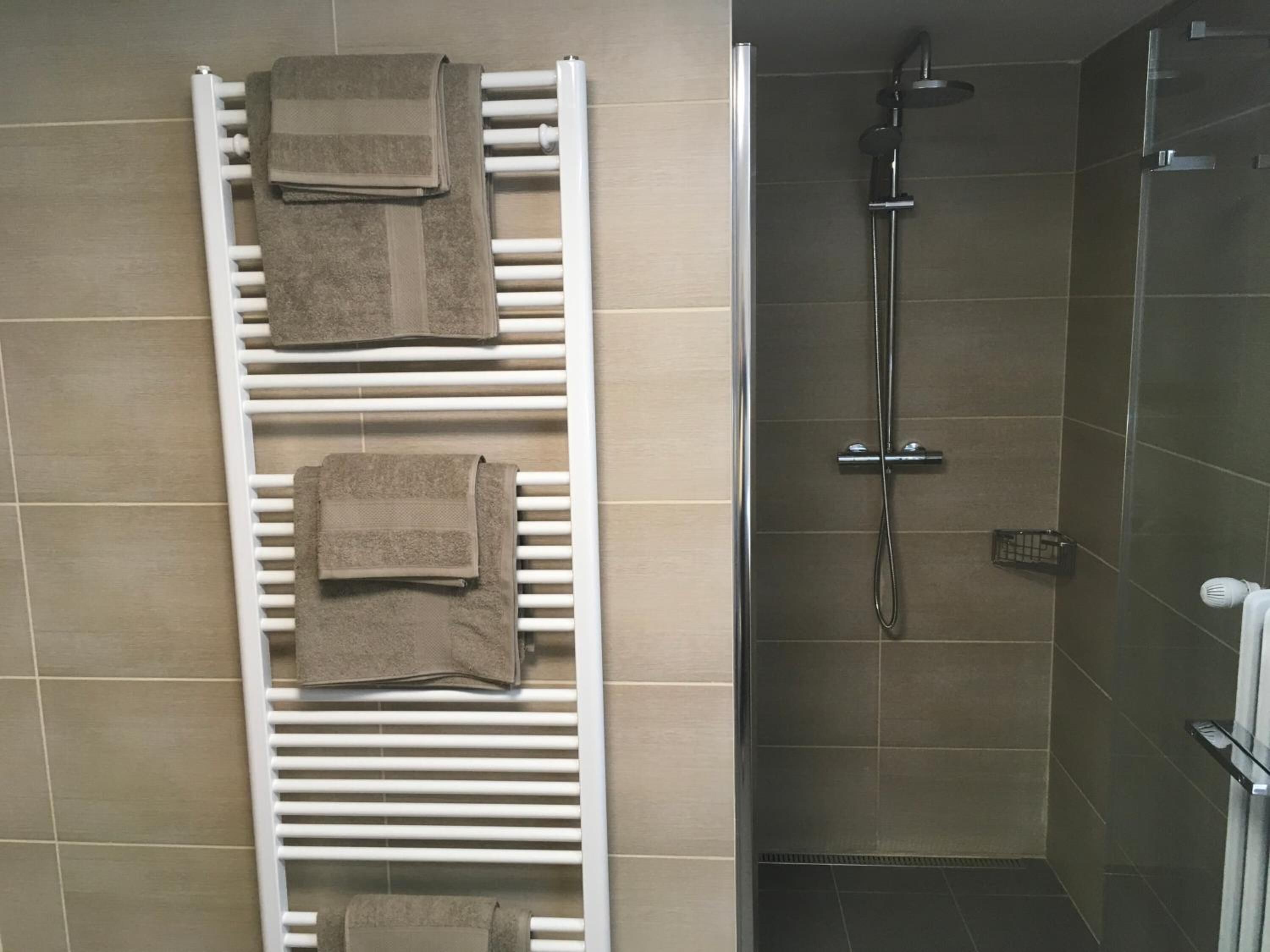Un baño con un perchero blanco que sostiene tres toallas, una zona de ducha con cabezal de ducha de mano y piso de baldosas.