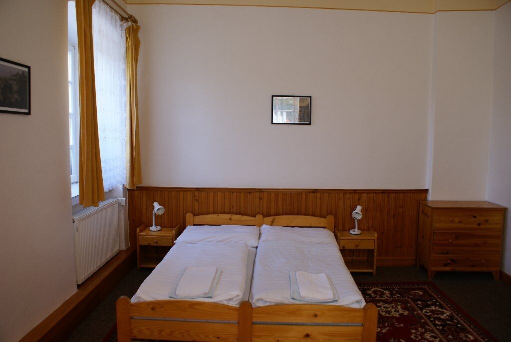 A double bed with two bedside tables, a small mirror, and a wooden dresser in a simple room with a radiator and a window with curtains.