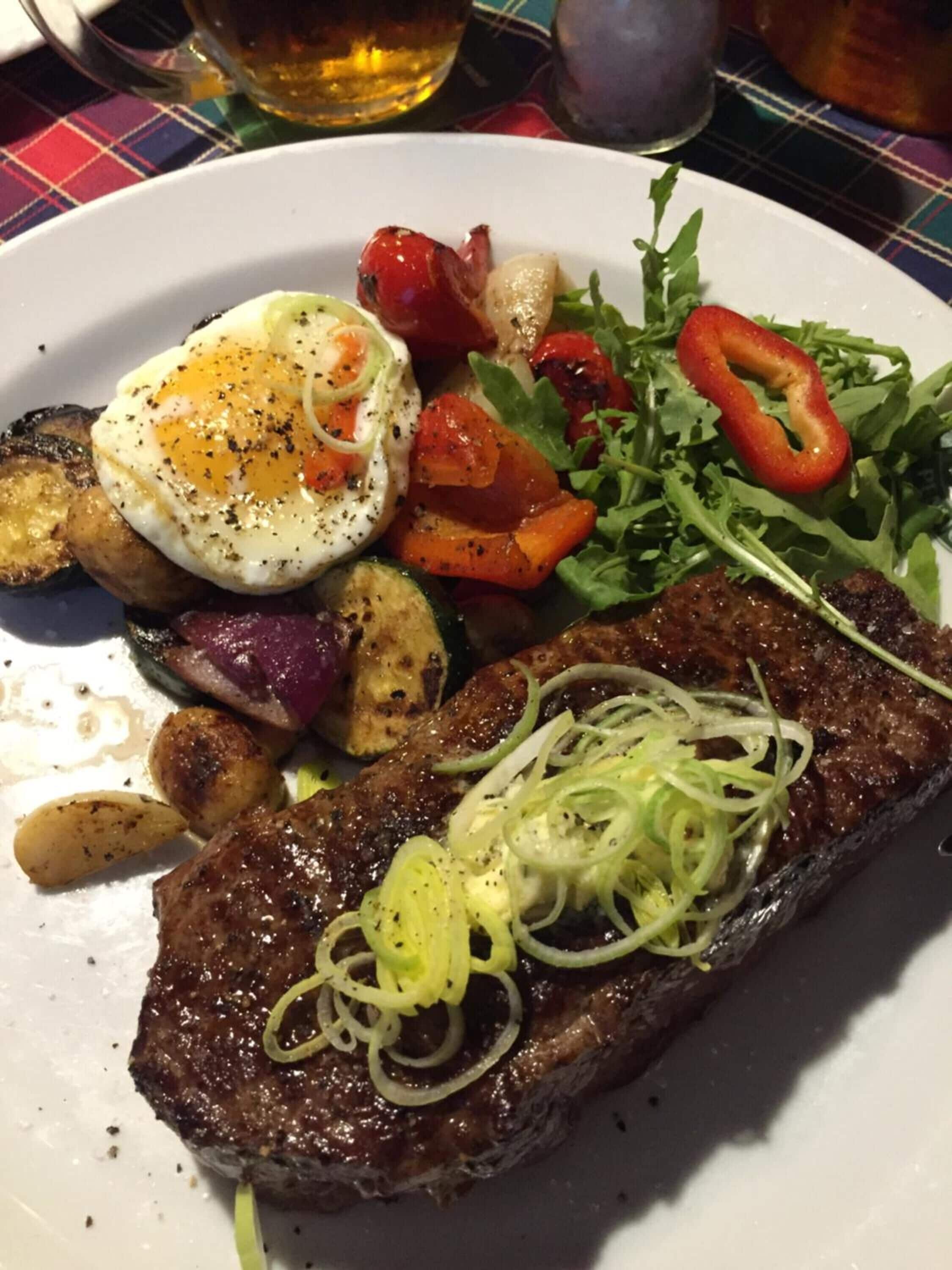 A plate with a steak topped with onion rings, a poached egg, roasted vegetables, and a side salad.