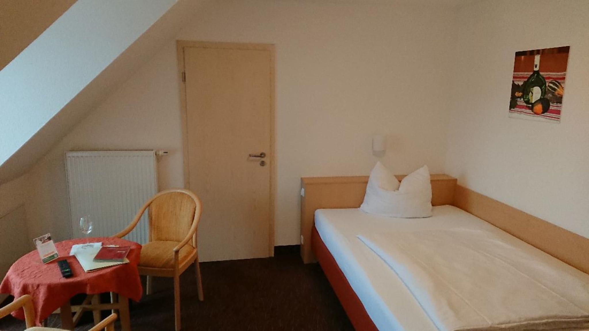 Ein Einzelbett mit Kissen, ein Holzhocker, ein kleiner Tisch mit roter Tischdecke und ein Heizkörper.