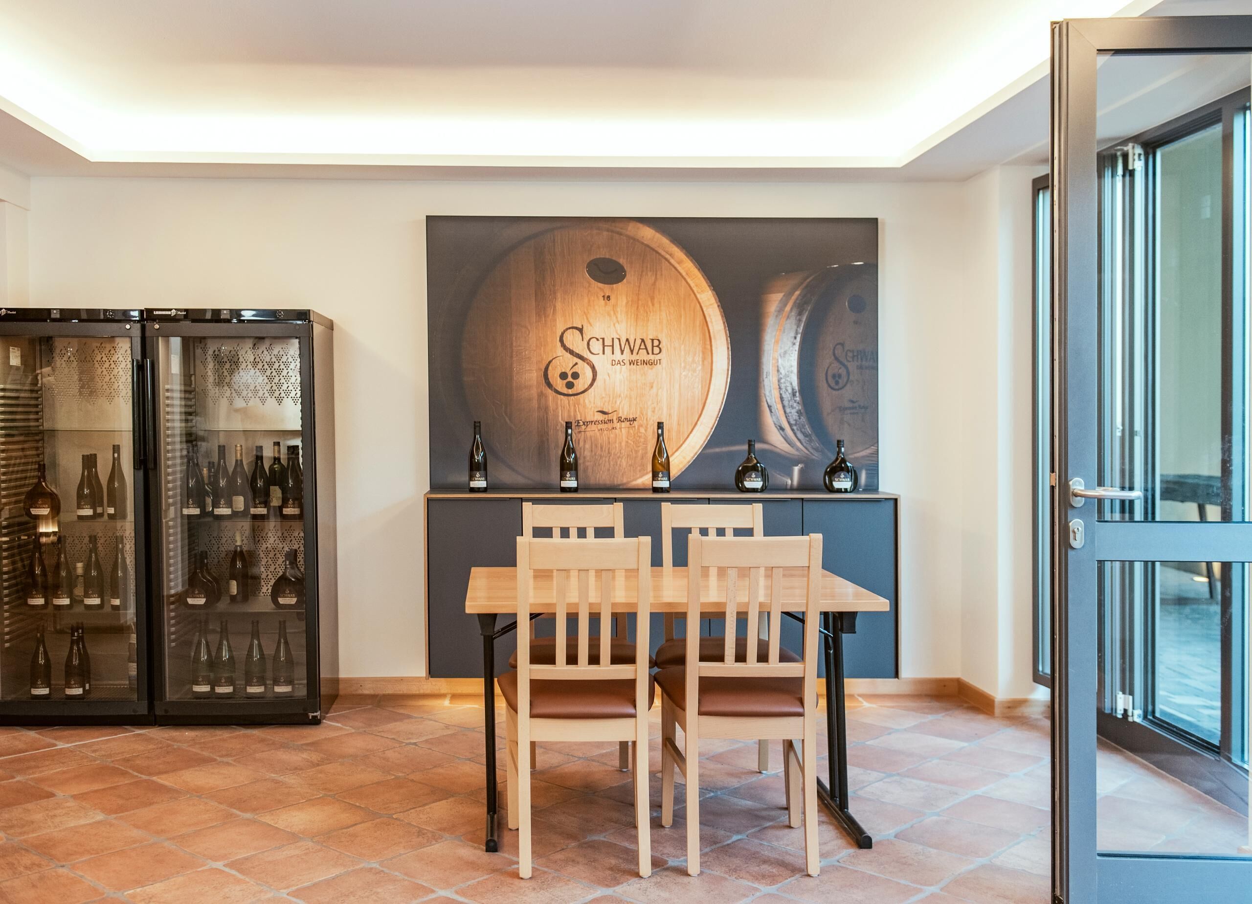 A dining area with a wooden table and chairs, a wine refrigerator, and a large wall-mounted wine barrel artwork.