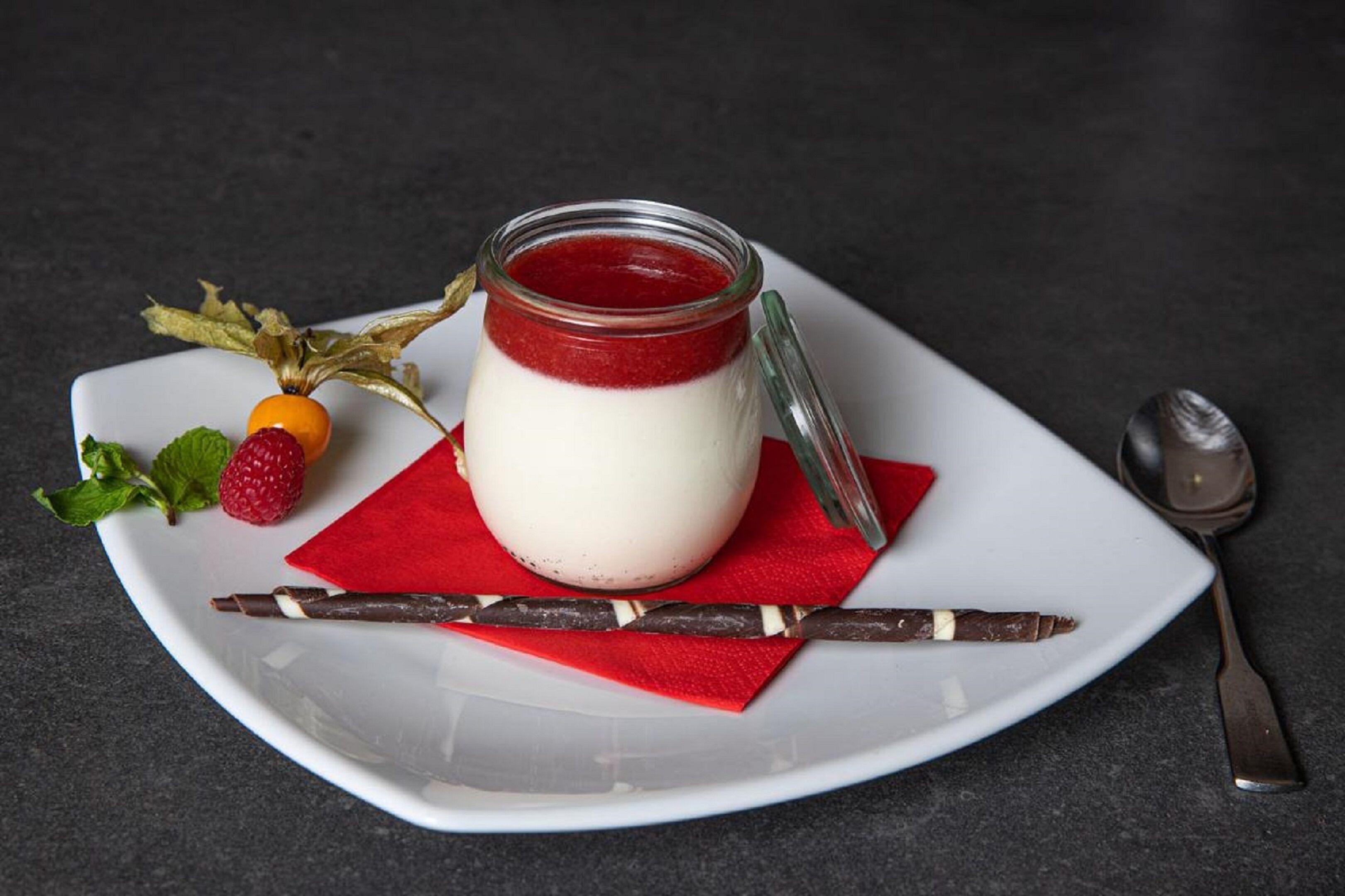 A dessert with a red jelly layer, a white creamy layer, and a chocolate stick on a white plate with a red napkin.