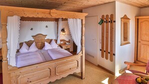 Una cama tradicional de cuatro postes de madera con sábanas blancas, una mesita de noche de madera, un sillón con alfombra roja, y una mesa con un cuenco de fruta y vasos.