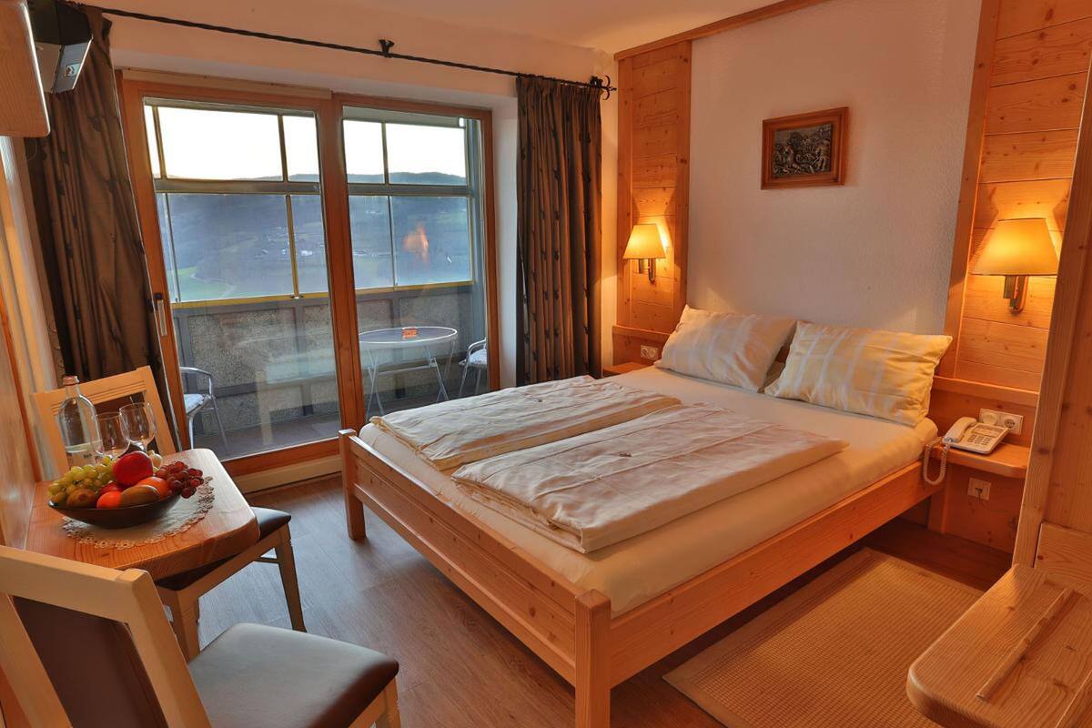 A bedroom with a wooden bed, a bedside table with a lamp, a small table with fruit, and a view of the outdoors through a sliding door.