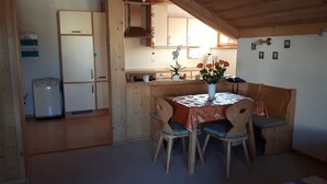 A compact kitchen with wooden cabinets, a small refrigerator, and a dining area with a table and chairs. - Ferienwohnung Bücherl (Waldmuenchen)