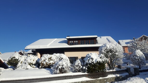 A snow-covered building with a sloped roof, surrounded by snow-laden trees and a stone wall. - Ferienwohnung Bücherl (Waldmuenchen)