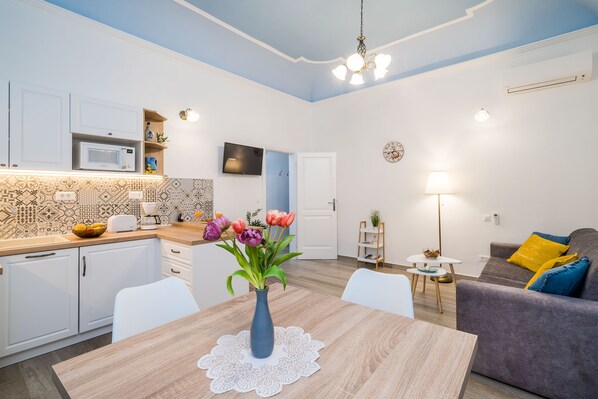 A modern kitchen with white cabinets, a microwave, and a coffee maker. There is a dining table with a vase of flowers. A gray sofa with blue and yellow pillows is in the living area.