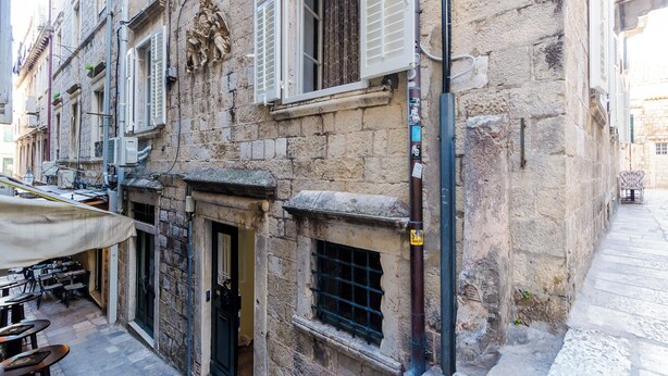A stone building with a door and windows, a small outdoor seating area with tables and chairs, and a narrow street. - Old city top spot apartments just one step from the main street Stradun (Dubrovnik)