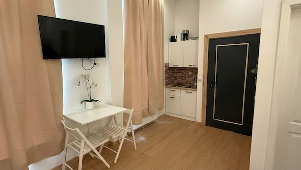 A compact room with a TV, a small white desk, a potted orchid, a folding chair, a kitchen area with a brick backsplash, and a black door.