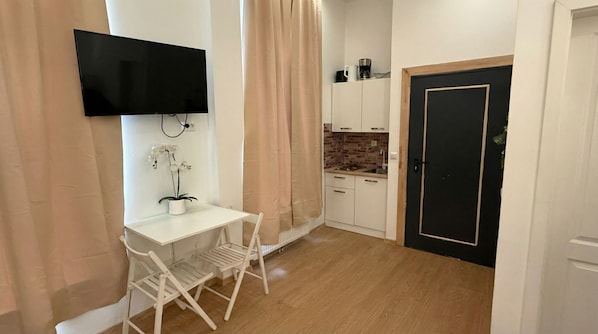 Quarto compacto com televisão, pequena escrivaninha branca, orquídea em vaso, cadeira dobrável, área de cozinha com painel de ladrilhos e porta preta.