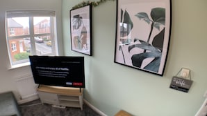 A room with a large window, a flat-screen TV on a stand, framed artwork on the wall, and a small wooden table. - The coach house views netflix sleeps 5 (Leicester)