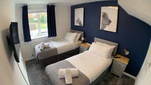 A hotel room with two single beds, a TV, a desk, and a window with curtains. - The coach house views netflix sleeps 5 (Leicester)