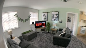 A living room with a grey sofa, a TV displaying '5 Reasons to Try Netflix', a small table with a lamp, and a kitchen area in the background. - The coach house views netflix sleeps 5 (Leicester)