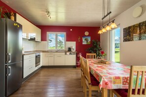 Uma cozinha moderna com frigorífico, forno e máquina de lavar loiça. Há uma mesa de jantar com cadeiras e toalha de mesa vermelha.