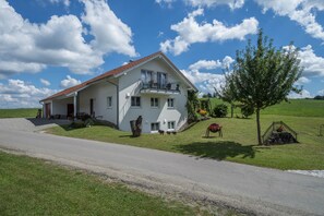 Uma casa de dois andares com varanda, entrada de autom óveis e gramado com árvore e canteiro de flores.