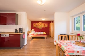 Ein kompakter Wohnbereich mit Küchenzeile, einem Bett mit bunter Bettwäsche und einer Essecke mit Tisch für zwei Personen.