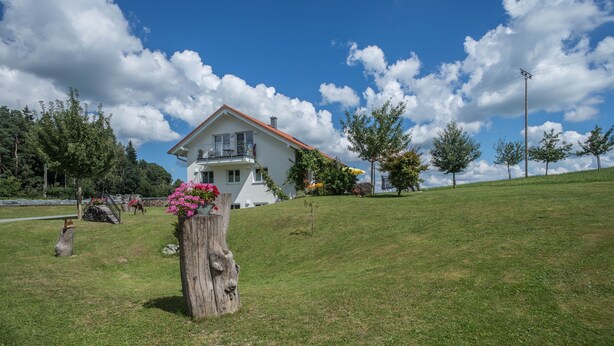 Une maison blanche avec un toit rouge, entourée d’une colline verdoyante et d’arbres.