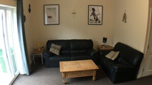 Ein Wohnzimmer mit einer schwarzen Ledersofa, einem Holztisch und einem kleinen Beistelltisch. An den Wänden hängen Bilderrahmen und es gibt eine Deckenleuchte.