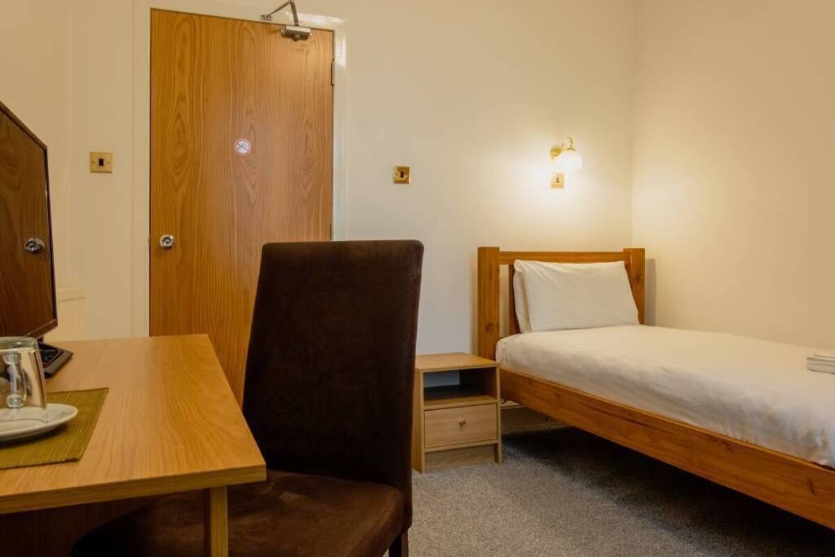 Una habitación pequeña y ordenada con un escritorio de madera, una silla, una cama individual, una mesita de noche y una lámpara en la pared.