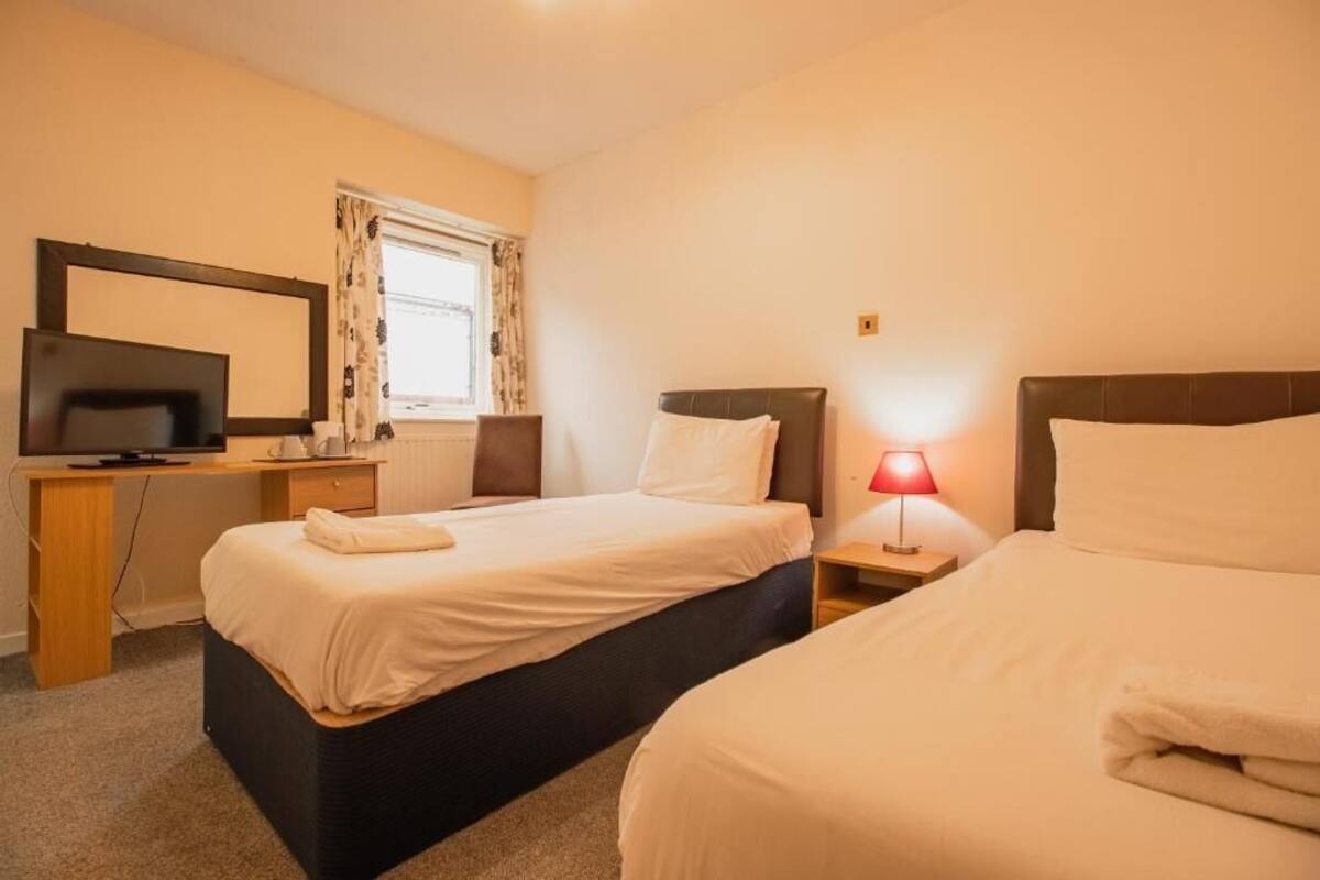Habitación de hotel con dos camas, un escritorio con televisor, una lámpara y una ventana con cortinas.